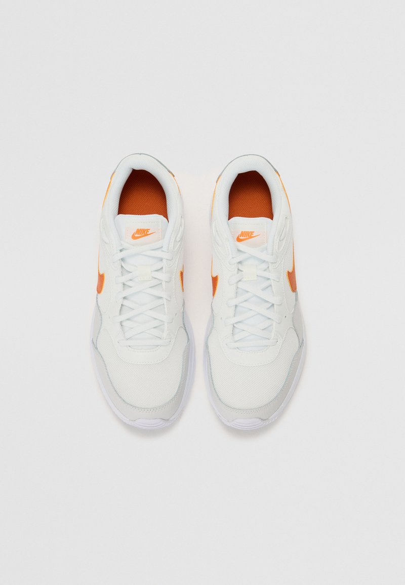 Zapatos deportivos blancos con acentos naranjas, tela texturizada, diseño con cordones y suela acolchada, con el logo de Nike en la lengüeta.
