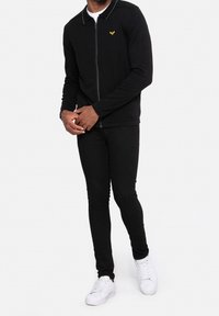 Homme portant un sweat à fermeture éclair noir avec un emblème jaune, un pantalon noir slim et des baskets blanches, debout devant un fond blanc.