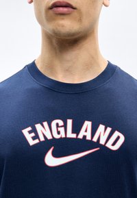 Κοντινό πλάνο ενός ατόμου που φοράει μπλε σκούρο πουκάμισο με τη λέξη "ENGLAND" και το λευκό λογότυπο Nike στη θωρακική περιοχή.