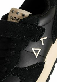 Sneaker nera realizzata con una combinazione di materiali in pelle e suede. Elementi di design notevoli includono un'etichetta beige, accenti color sabbia e superfici testurizzate.