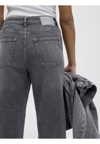 Grå denimjeans med avslappnad passform, med bakfickor och en texturerad yta; tillsammans med en matchande grå jacka som hålls i handen.