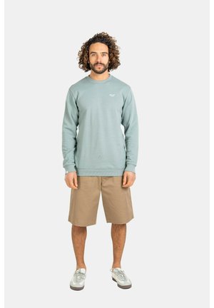 Mann mit lockigem Haar, der einen hellblauen Pullover, beige knielange Shorts und weiße Turnschuhe trägt, steht vor einem einfarbigen Hintergrund.