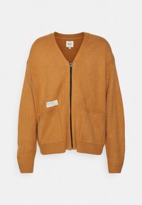 Orange stickad cardigan med v-ringning, dragkedja framtill och två stora påsydda fickor. Har en etikett på den vänstra fickan.