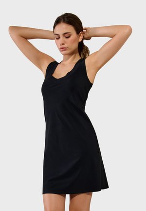 Mujer con pelo castaño, vestida con un vestido negro sin mangas, posando con los brazos levantados y los ojos cerrados sobre un fondo claro liso.
