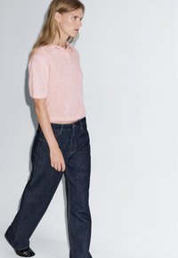 Světle růžový pletený polo svetr s krátkými rukávy, doplněný tmavými džínami širokých nohavic. Jednoduchý design s jemnou texturou a detaily.