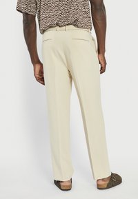 Pantalon beige avec une coupe ajustée, doté de deux poches arrière et d'une texture lisse. Porté avec des sabots marron, présenté de dos.