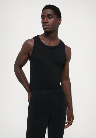Joven con una camiseta sin mangas negra y pantalones negros, de pie con una mano detrás de la espalda, mirando de lado.