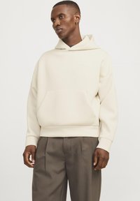 Lichte crèmekleurige hoodie met een voorzak, ribgebreide manchetten en zoom. Soepele textuur, relaxte pasvorm, gecombineerd met donkere, op maat gemaakte broeken.