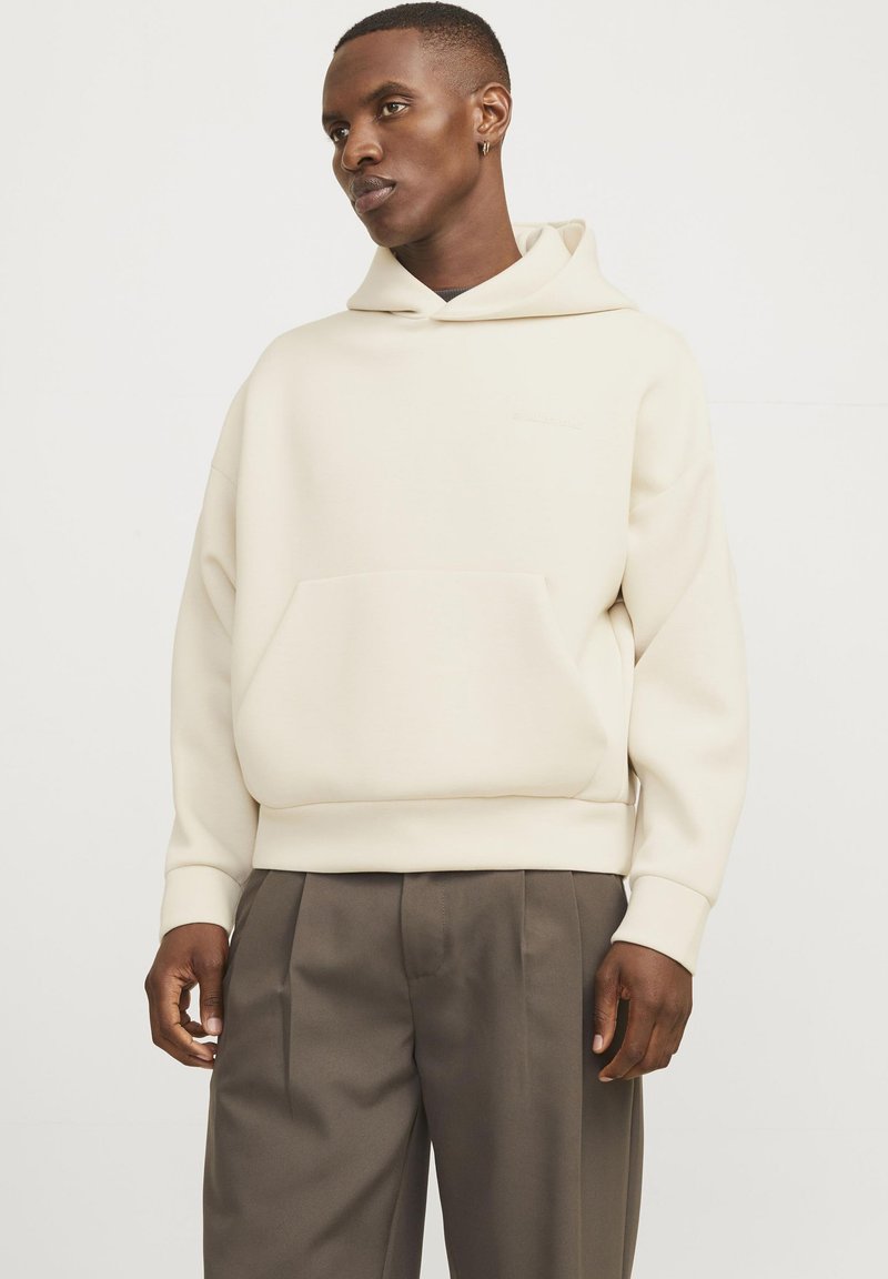 Lichte crèmekleurige hoodie met een voorzak, ribgebreide manchetten en zoom. Soepele textuur, relaxte pasvorm, gecombineerd met donkere, op maat gemaakte broeken.