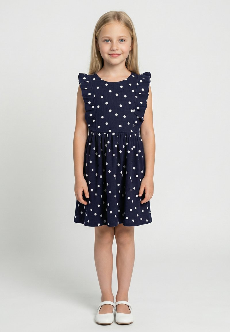 Granatowa sukienka w białe groszki, z falbanami na ramiączkach i elastyczną talią. Do tego białe baleriny.