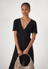 Robe noire à manches courtes avec un décolleté en V profond et une fente à l'avant. Portée avec un collier circulaire en argent et un sac à main en peau de serpent beige.