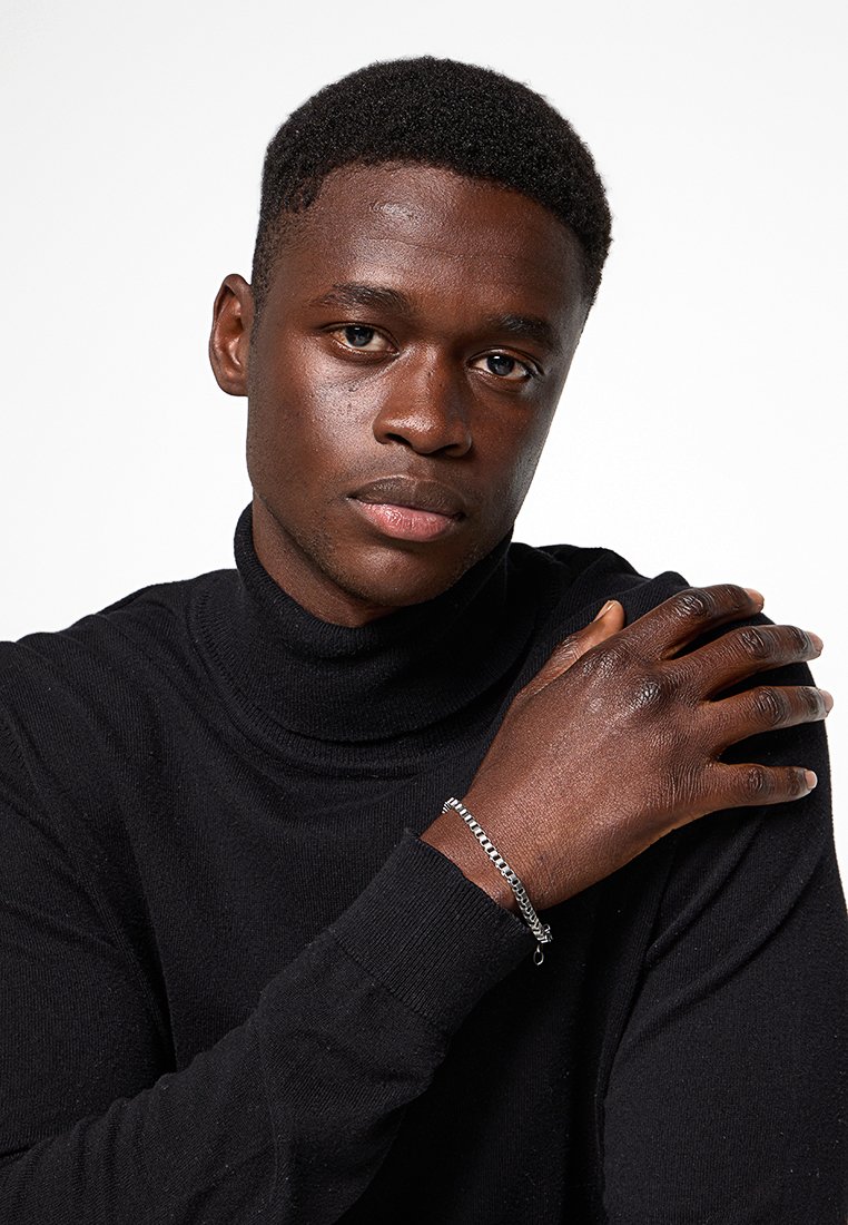 Silberne Kettenarmband, das an einem Handgelenk mit dunkler Haut getragen wird, mit ineinandergreifenden Gliedern und einem minimalistischen Design vor einem schwarzen Rollkragenpullover.