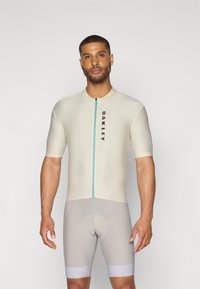 Combinaison de cyclisme couleur crème avec des manches courtes, une fermeture éclair à l'avant en turquoise et un logo noir. Comprend un short de cyclisme rembourré gris.