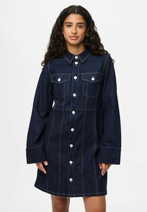 Frau in einem dunkelblauen, knöpfigen Jeanskleid mit langen Ärmeln und Brusttaschen, steht vor einem weißen Hintergrund.