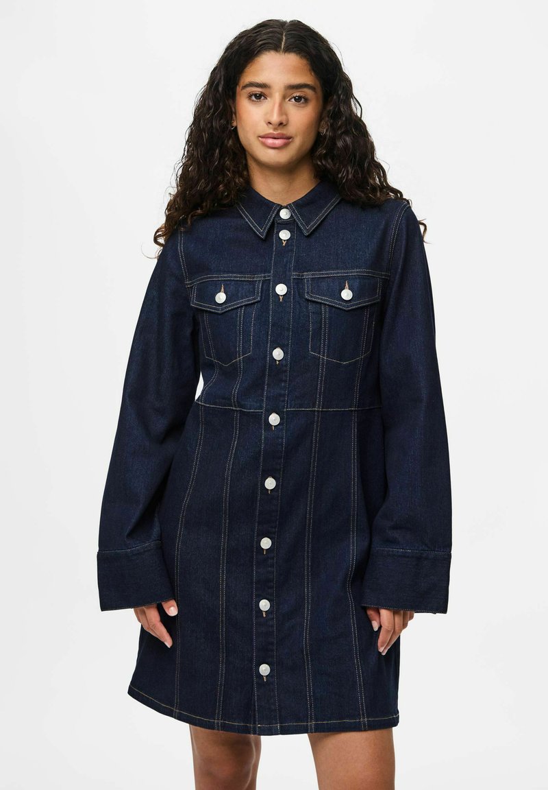 Femme portant une robe en denim bleu foncé à boutons avec des manches longues et des poches poitrine, se tenant contre un fond blanc.