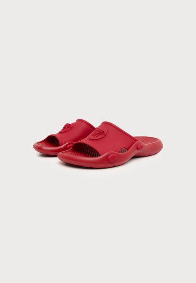 Paire de sandales rouges à enfiler avec orteils ouverts et semelles texturées, présentant un emblème de lion en relief sur les brides.