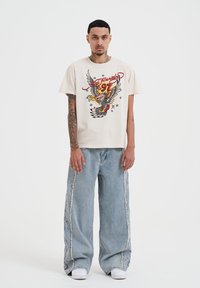 Béžové grafické tričko s designem orla a číslem 97, spárované se světle modrými širokými džíny s třásněmi a bílými teniskami.