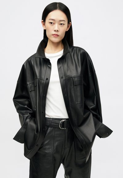 Νέος άνθρωπος με μακριά μαύρα μαλλιά, φορώντας μαύρο υπερμεγέθες δερμάτινο μπουφάν και παντελόνια, λευκό πουκάμισο, ποζάροντας με τα χέρια στις τσέπες του μπροστά σε λευκό φόντο.