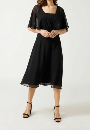 Kobieta w czarnej sukience do kolan z przezroczystymi falbanami na rękawach i czarnych sandałach na wysokim obcasie z paskiem wokół kostki, stojąca z rękami splecionymi.