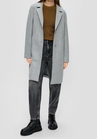 Cappotto grigio in misto lana con revers a punta, due bottoni, indossato su un maglione marrone a coste e jeans neri a vita alta, abbinato a stivali neri.
