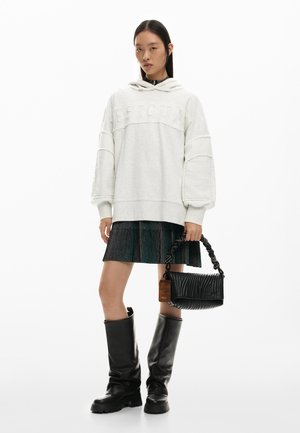 Joven mujer con sudadera blanca de gran tamaño, falda corta negra plisada, botas negras hasta la rodilla, sosteniendo un bolso negro con una correa texturizada.