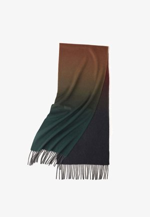 Olivgrüner Pullover, beiger Hosen und weiße Sneakers. Ein mehrfarbiger Schal liegt um den Hals und hat Fransen. Eine schwarze Handtasche in der Hand.