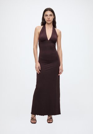 Mujer que lleva un vestido de tirantes sin mangas de color marrón oscuro con cuello halter y sandalias de tiras, de pie frente a un fondo claro y liso.