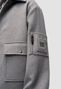 Šedá vlněná bunda s límcem, velkými předními kapsami a kapsou na zip na rukávu s vytlačeným logem značky. Měkká textilní struktura.