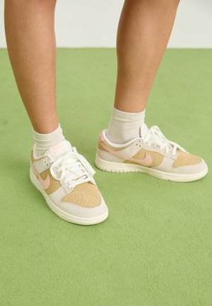 Jambes portant des chaussettes blanches et des baskets Nike beiges et roses debout sur un sol recouvert de moquette verte.