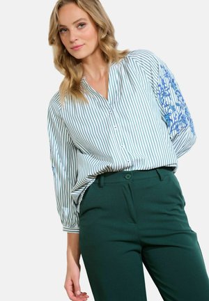 Camicia con bottoni, a strisce verticali verdi e bianche, con ricamo blu sulla manica sinistra. Abbinata a pantaloni verde scuro.