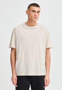 Beige puuvillainen t-paita lyhyillä hihoilla, pyöreällä kaula-aukolla ja hienovaraisella pyöreällä logolla rinnassa. Sujuva kangas, rento istuvuus.