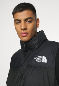 Black insulated vest with high collar, zip closure, and logo; features a smooth texture and panel design. Suitable for cold weather.