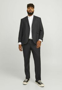 Costume noir avec une veste à simple boutonnage, pantalon assorti, chemise blanche et baskets noires. Tissu texturé, coupe ajustée et sans motifs visibles.
