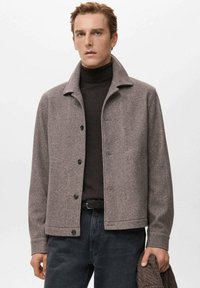 Chaqueta de lana gris con cuello clásico, botones delanteros y dos bolsillos laterales, combinada con un suéter de cuello alto oscuro y vaqueros oscuros.