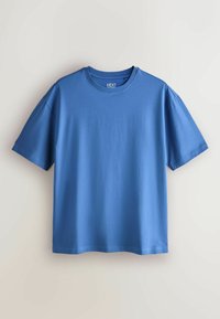 Lihtne sinine lühikeste varrukatega ümara kaelusega T-särk ripub heleda beeži seina taustal, krae sees olev silt kannab teksti "NEXT".