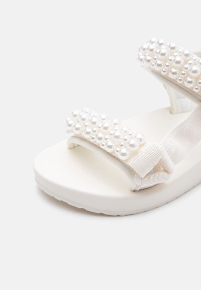 Λευκό σανδάλι με δύο λουράκια διακοσμημένα με συστάδες από λευκές ψεύτικες πέρλες και μαλακή σόλα.