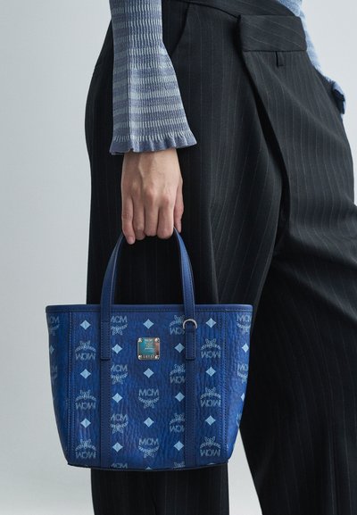 Χέρι κρατάει μπλε τσάντα MCM με λογότυπο, φορώντας καρό μπλούζα με μακριά μανίκια και μαύρο παντελόνι με ρίγες σε ανοιχτό φόντο.