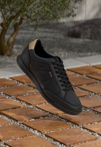 Black sneaker featuring a smooth leather upper, a gold-accented heel, and a textured rubber sole, displayed above a patterned tile surface.