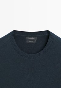 Ciemnoniebieski sweter bawełniany z okrągłym dekoltem, prążkowanym kołnierzem i ściągaczami, z metką marki naszytą wewnątrz kołnierza.