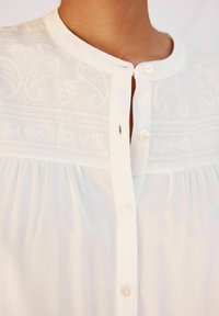 Weißes Bluse mit runder Ausschnitt, vorderem Knopfverschluss und aufwendigen Stickdetails. Weicher, leichter Stoff mit Falten.