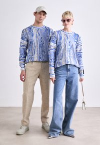 Des pulls bleu à motifs avec des designs texturés, assortis à un pantalon beige et un jean large bleu clair. Les accessoires comprennent une casquette et des lunettes de soleil.