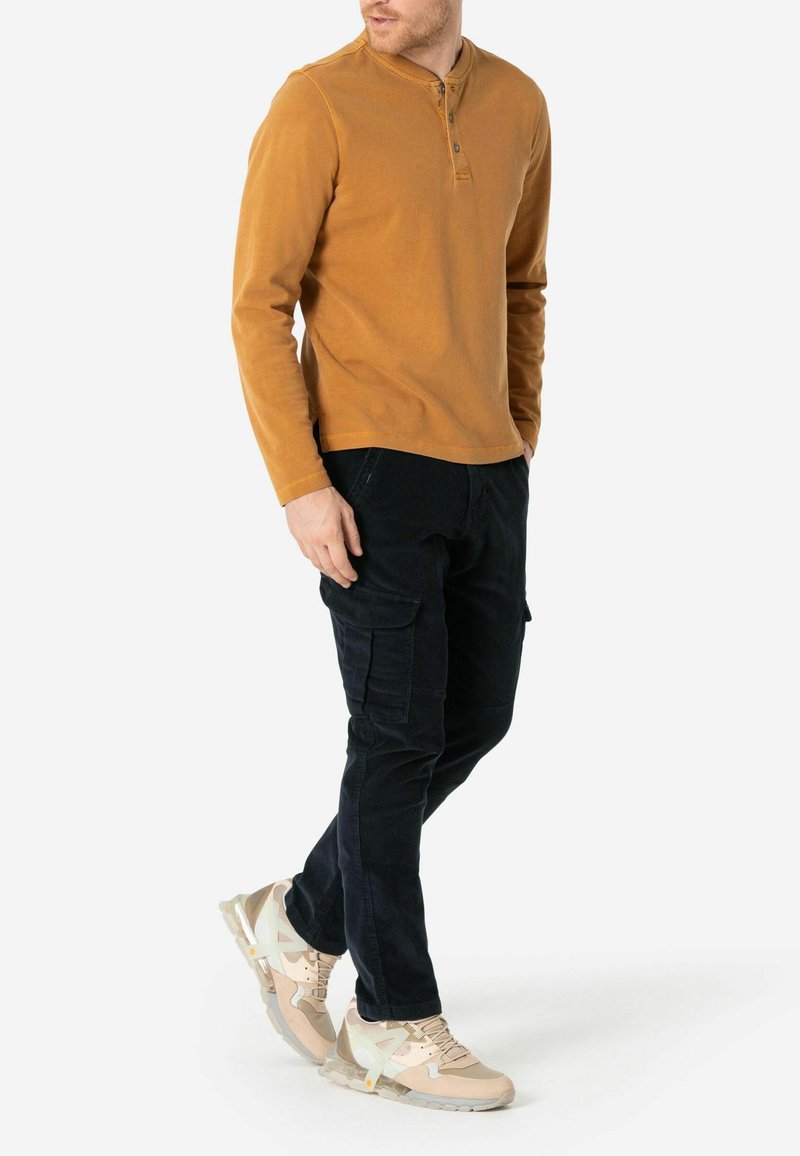 Langärmliges, senfgelbes Henley-Shirt mit rundem Ausschnitt und Knöpfen, kombiniert mit schwarzen Cargo-Hosen und hellbeigen Sneakers.