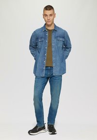 Camisa de denim azul con dos bolsillos en el pecho, combinada con una camiseta verde oliva y jeans azules, terminada con zapatillas negras.