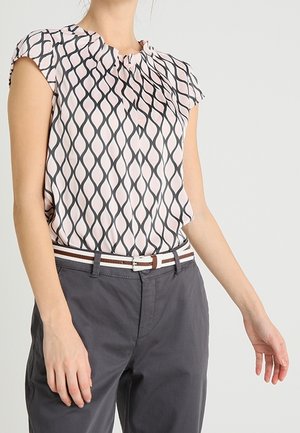 Persoon met een korte mouw patroon blouse die ingestopt is in donkergrijze broek met een wit en bruin gestreepte riem tegen een effen achtergrond.