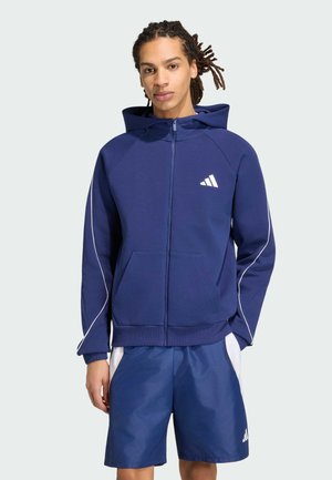 Ung mand med dreadlocks iført en mørkeblå hættetrøje med lynlås og hvidt logo samt matchende shorts med hvide sidepaneler.