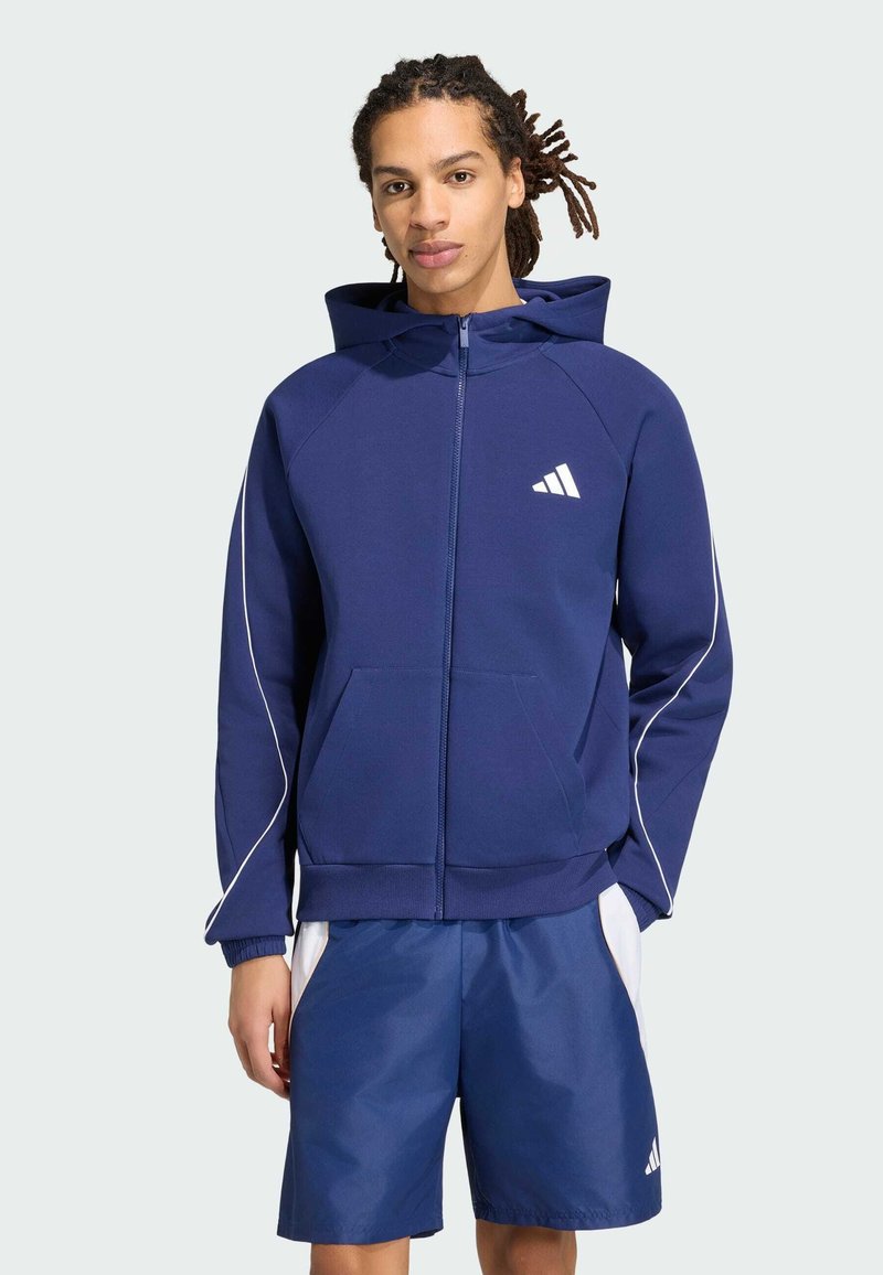 Jeune homme avec des dreadlocks portant un sweat à capuche zippé bleu marine avec un logo blanc et un short assorti avec des panneaux latéraux blancs.