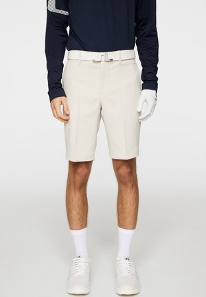 Beige golfshorts met voorplooien, een witte riem en een effen textuur. Gecombineerd met een marineblauwe langemouwen top en witte sportschoenen.