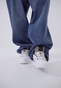 Baskets blanches avec des accents noirs, présentant une surface texturée et des lacets. Associées à un pantalon en denim bleu lâche légèrement oversize.