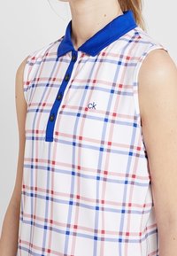 Ärmlös, kragklädd polo i vitt med ett rutigt mönster i blått, rött och marinblått. Har svarta knappar och en broderad "ck"-logotyp på bröstet.