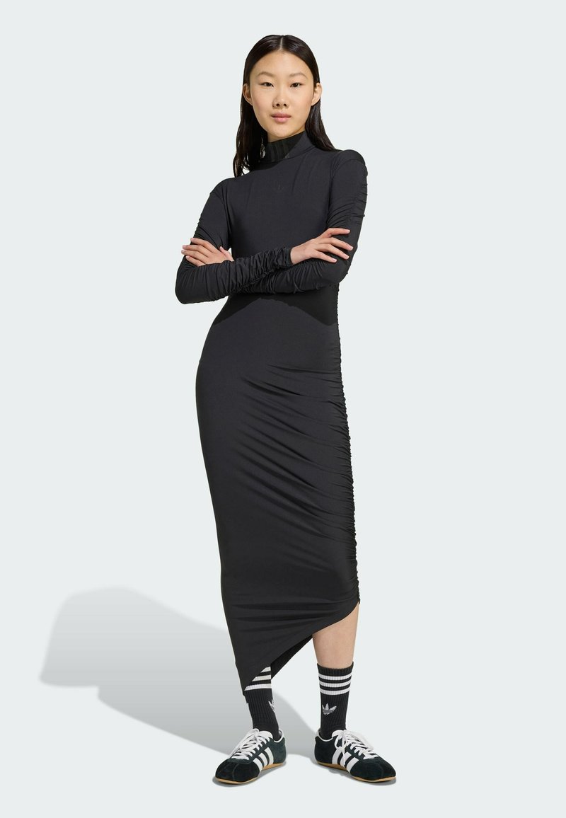 Svart långärmad klänning med hög krage; har rynkor på sidorna för textur. Kombineras med svart och vita träningsskor och strumpor.